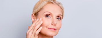 A mature woman with fair skin and blonde hair gently touching her face with one hand
